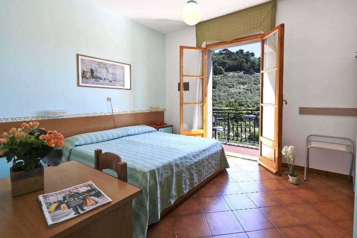 Un dormitorio con cama, un escritorio con revista, una silla, un ventanal con vista a la vegetación y un balcón con barandilla.