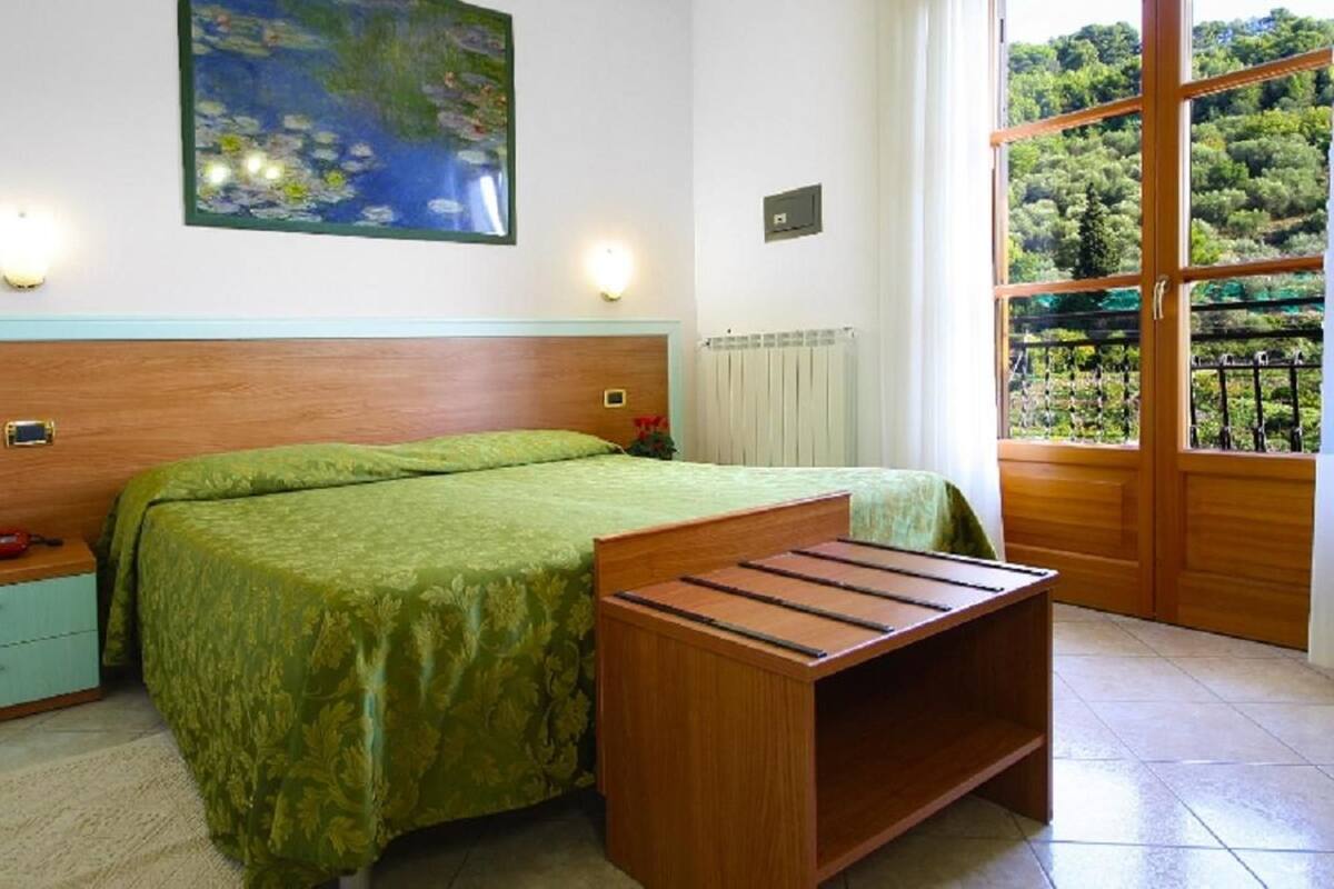 Un dormitorio con cama, mesita de noche, lámpara y vistas a la vegetación a través de una ventana.