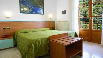 Een slaapkamer met een bed, nachtkastje, lamp en uitzicht op groen door een raam.