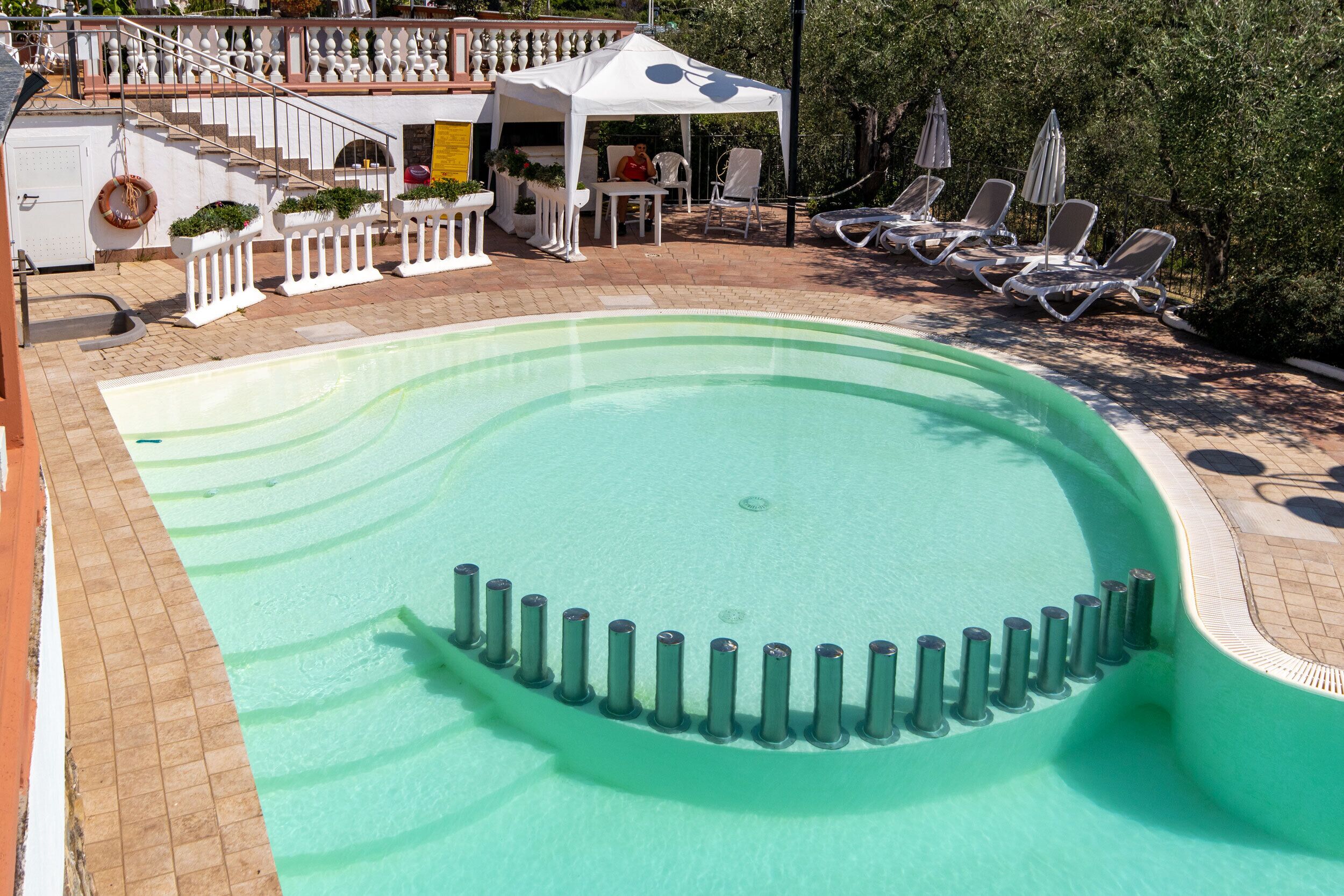 A swimming pool with a curved edge, surrounded by lounge chairs and a covered seating area with a railing.