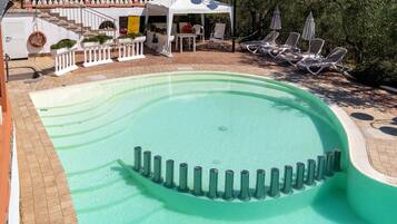 A swimming pool with a curved edge, surrounded by lounge chairs and a covered seating area with a railing.