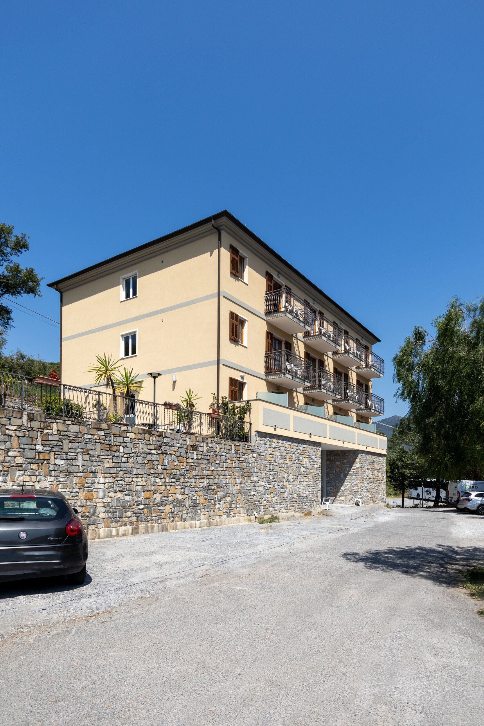 A multi-story building with balconies, a stone retaining wall, and a parking area.