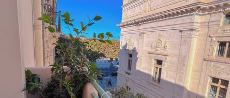 Un balcón con una maceta, baranda metálica y vista a un edificio histórico de arquitectura ornamentada.