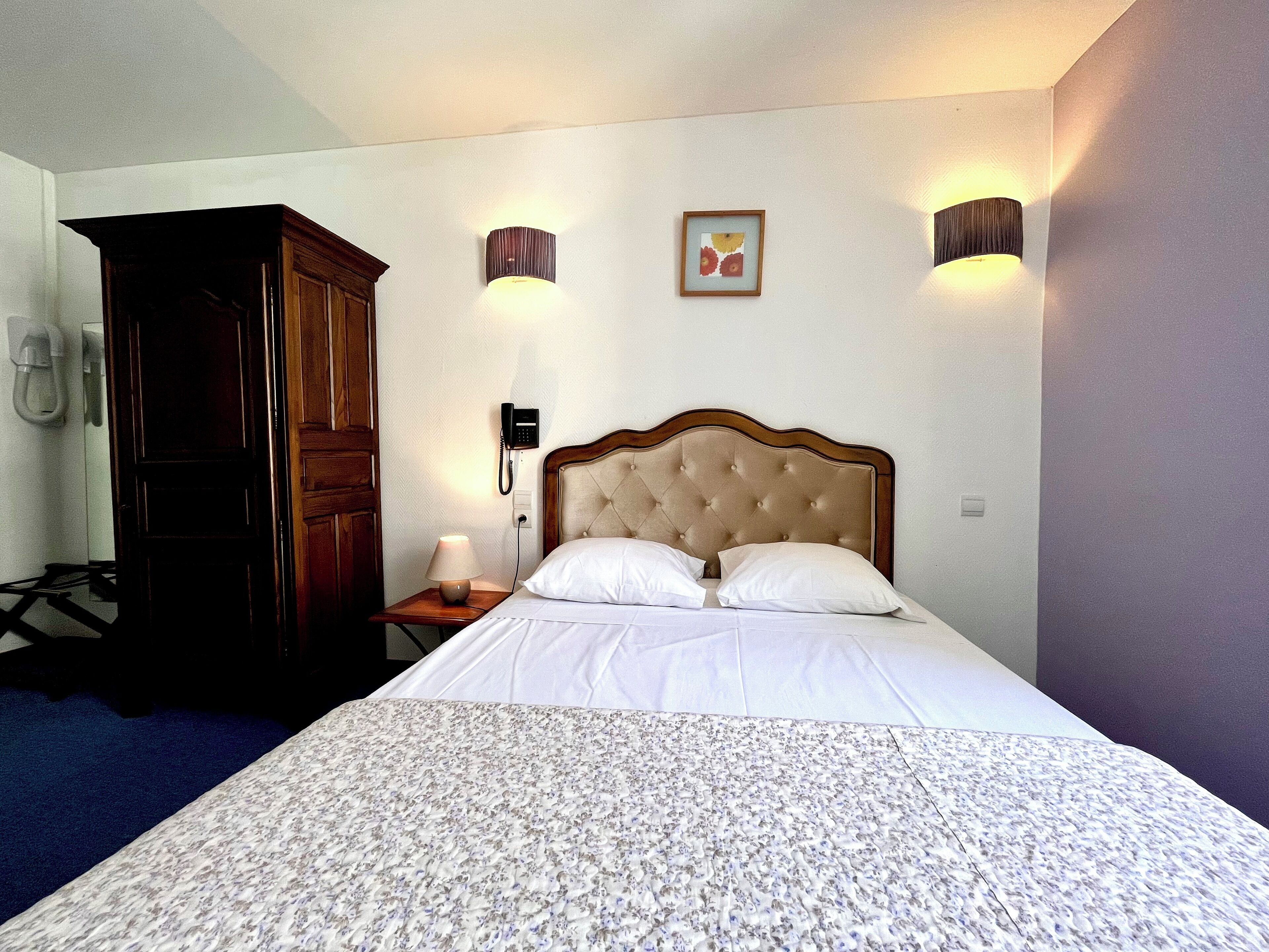 Habitación de hotel con cama, armario de madera, lámpara de noche, teléfono e iluminación en la pared.