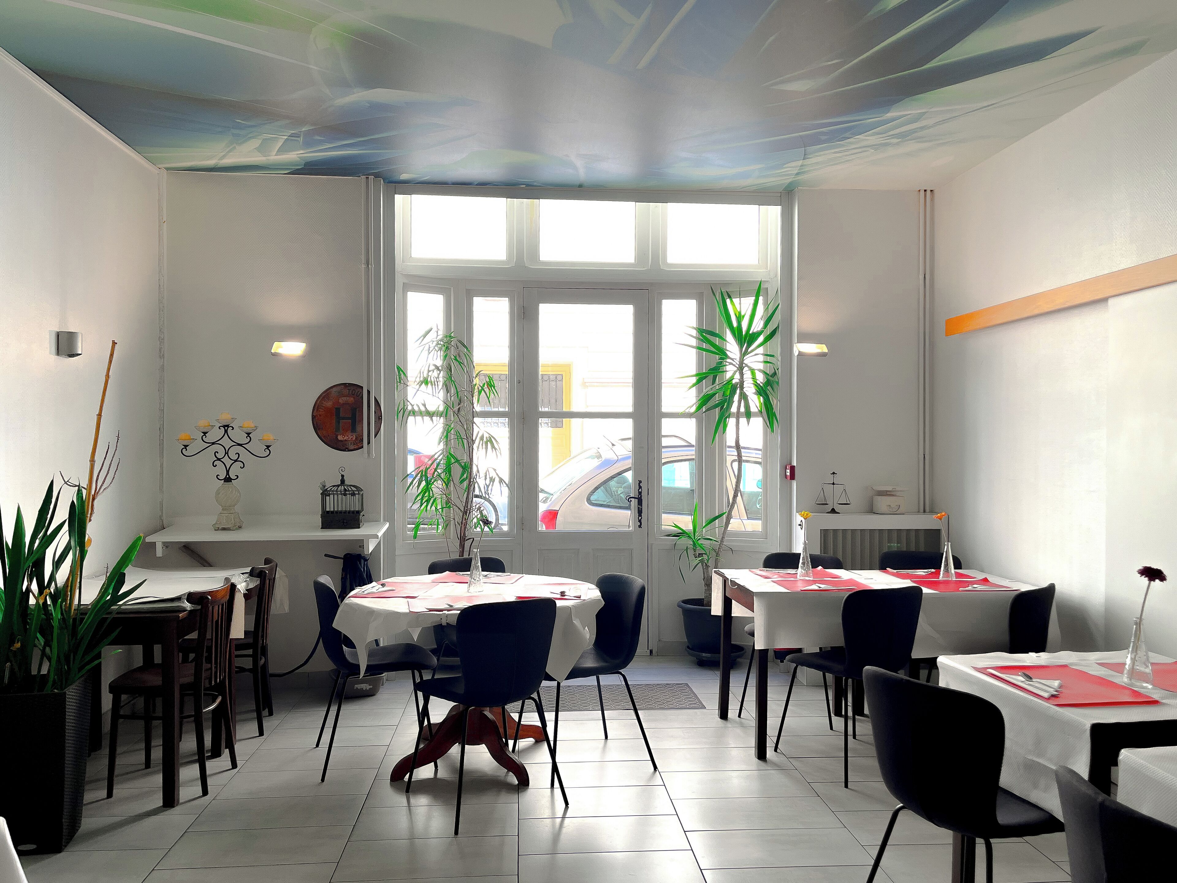 A restaurant with multiple tables and chairs, large windows, and indoor plants.