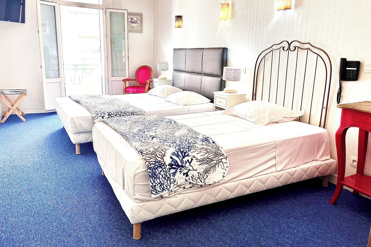 Una cama doble con cabecera acolchada, una mesita de noche con lámpara, una silla roja y un taburete de madera.