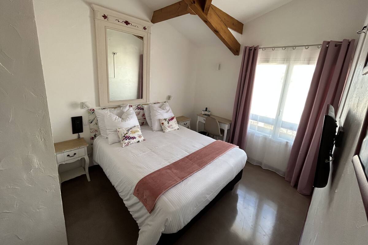 Un dormitorio con una cama grande, una viga de techo de madera, un espejo, un escritorio y una ventana con cortinas.