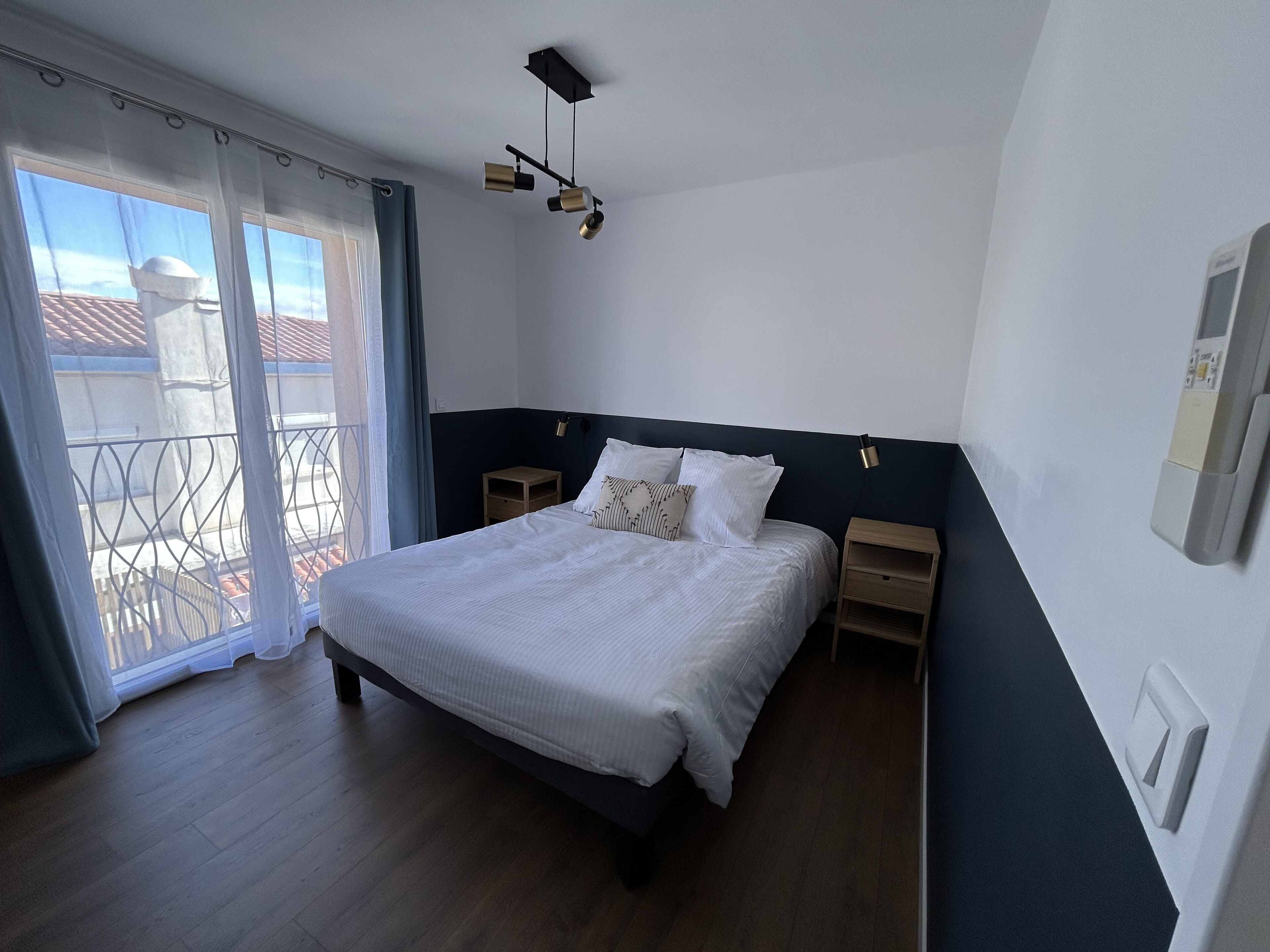 Ein Schlafzimmer mit einem Bett, einem Nachttisch, einer Wandleuchte und Blick durch ein Fenster mit Vorhängen.