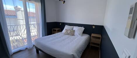 Ein Schlafzimmer mit einem Bett, einem Nachttisch, einer Wandleuchte und Blick durch ein Fenster mit Vorhängen.
