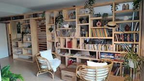 A cozy reading nook with two wicker chairs, a wooden bookshelf filled with books and decorative items, and a variety of potted plants.
