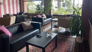 A modern living room with a black leather sofa, a coffee table, and a patterned rug. There are multiple seating options including a couch and chairs. Large windows provide a view of the outdoors. - Hotel Kristina (Imperia)