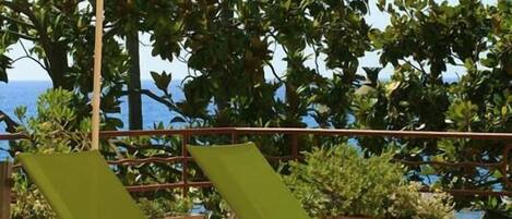 Terraza con dos sillones verdes y una sombrilla blanca, con vista a un cuerpo de agua y palmeras.