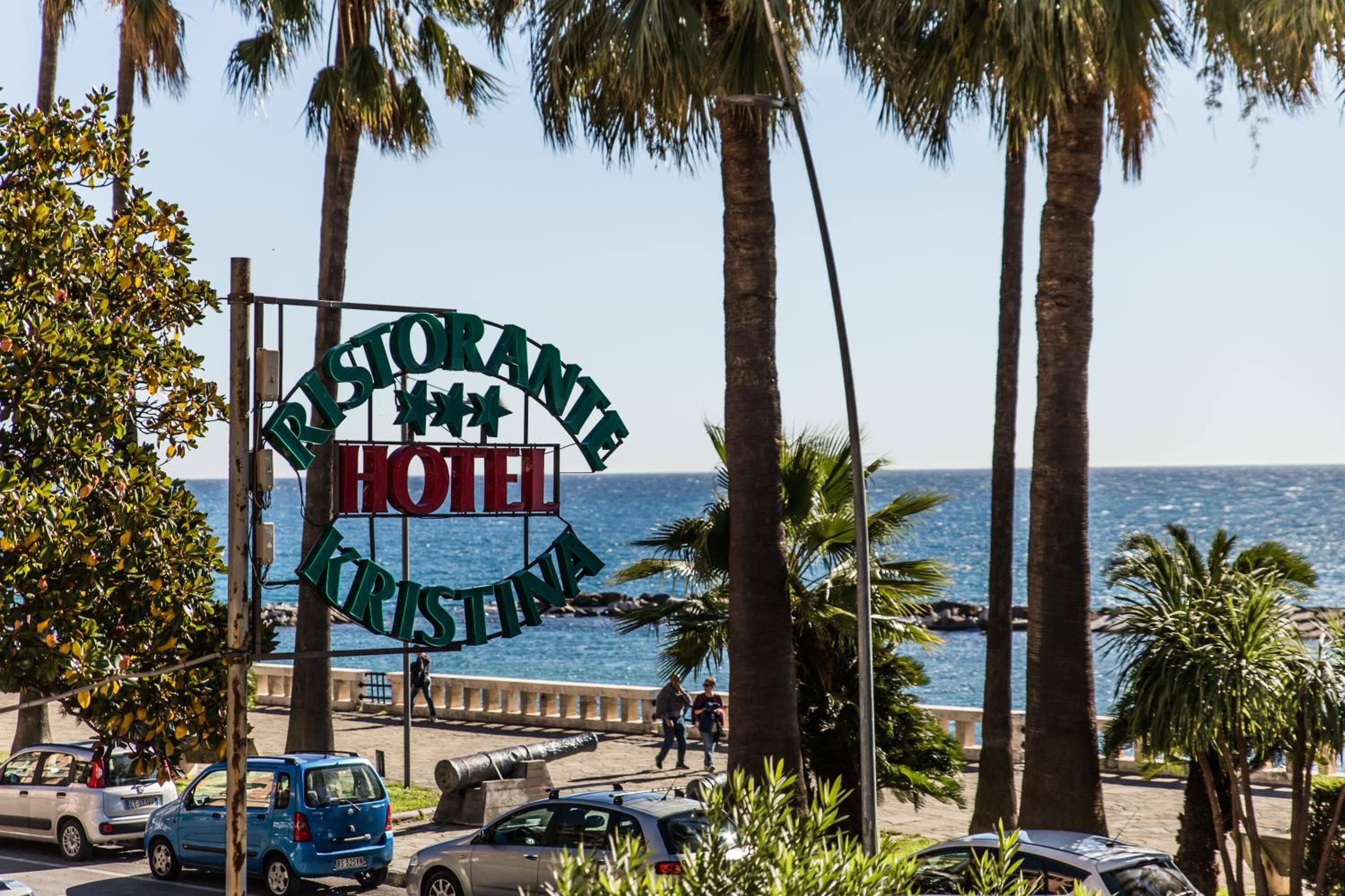 A sign for a hotel named "Ristorante Krisztina" with a view of the sea and palm trees.