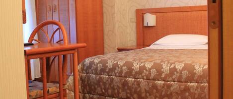 A bedroom with a wooden wardrobe, a bed with a floral bedspread, a wooden desk with a chair, and a patterned wallpaper.
