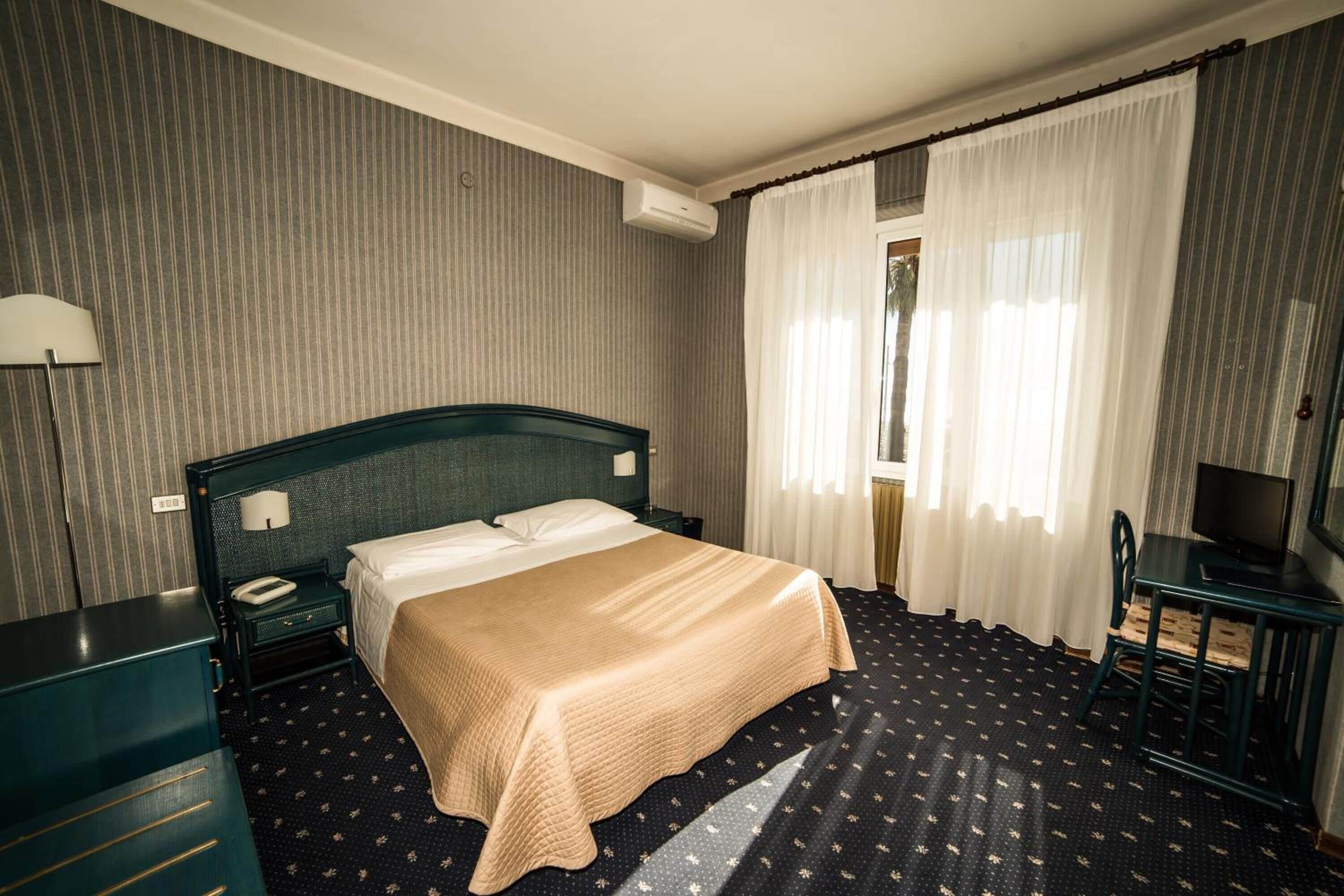 A hotel room with a bed, a desk, a chair, a lamp, and curtains.