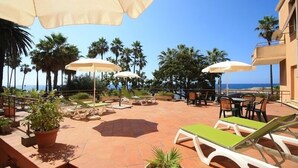 An outdoor patio with lounge chairs and umbrellas, surrounded by palm trees and a view of the ocean. - Hotel Kristina (Imperia)