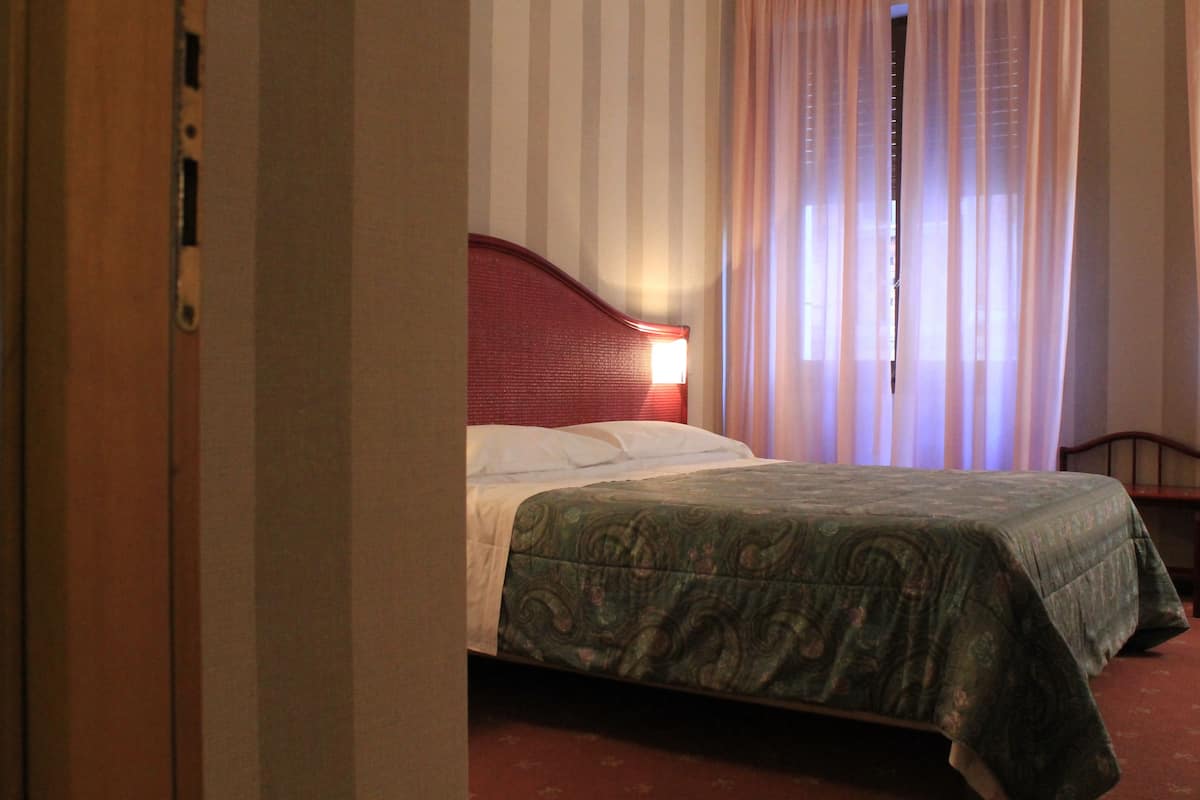 Ett hotellrum med en säng, en träpanel som huvudgavel, ett fönster med tunna, genomskinliga gardiner och en röd vägg.