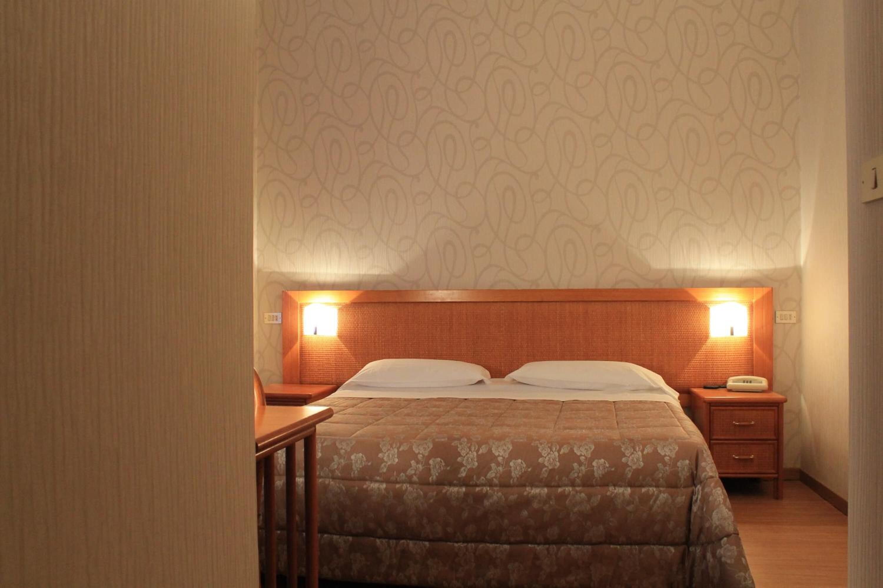 A hotel room with a wooden headboard, two bedside lamps, a bed with a floral-patterned bedspread, a nightstand with a telephone, and a wooden chair.