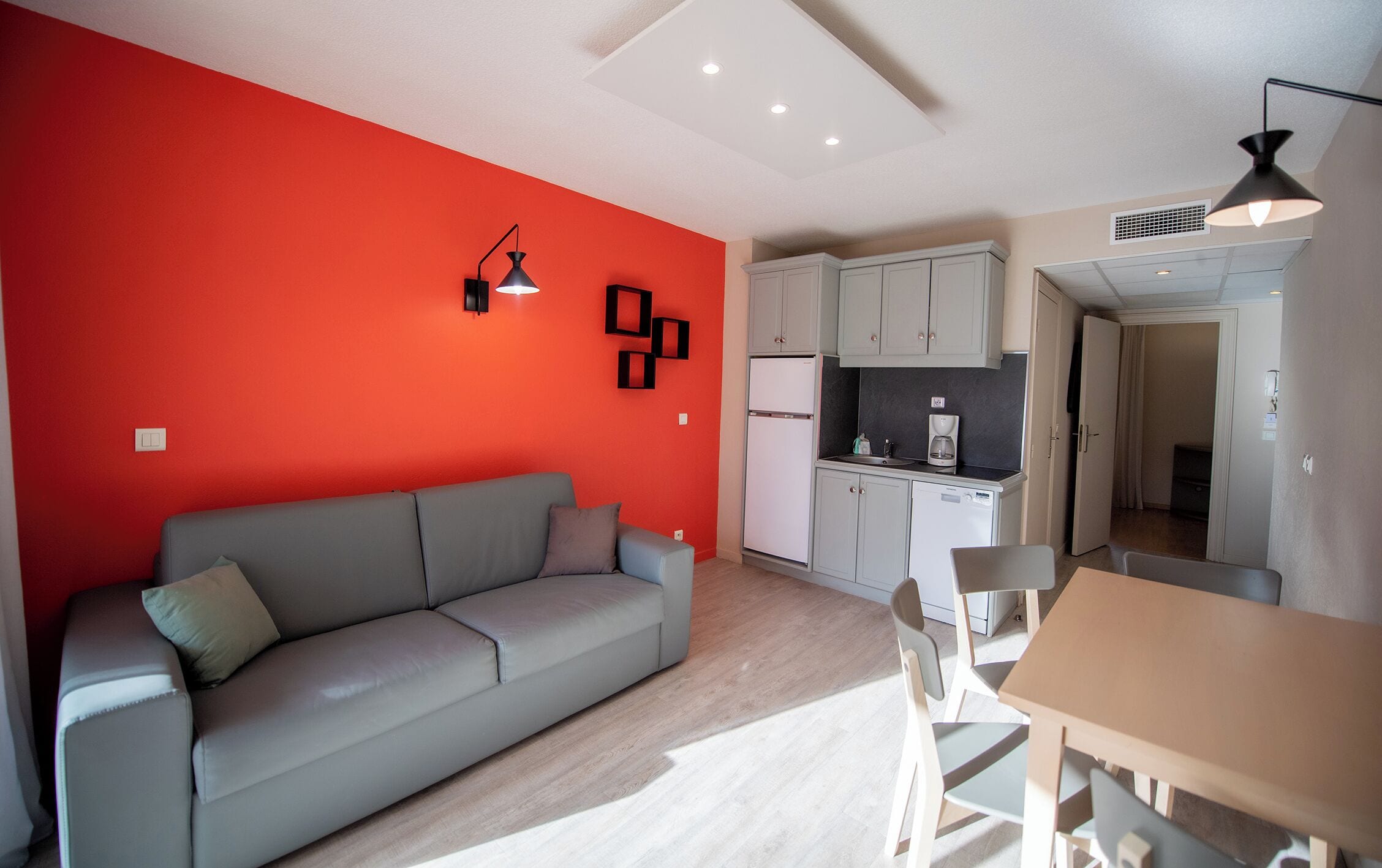Un espace de vie compact comprenant un canapé gris, une table à manger avec des chaises, un coin cuisine avec des placards et des appareils électroménagers, et un mur d’accent rouge.