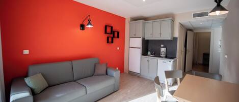 Un espace de vie compact comprenant un canapé gris, une table à manger avec des chaises, un coin cuisine avec des placards et des appareils électroménagers, et un mur d’accent rouge.