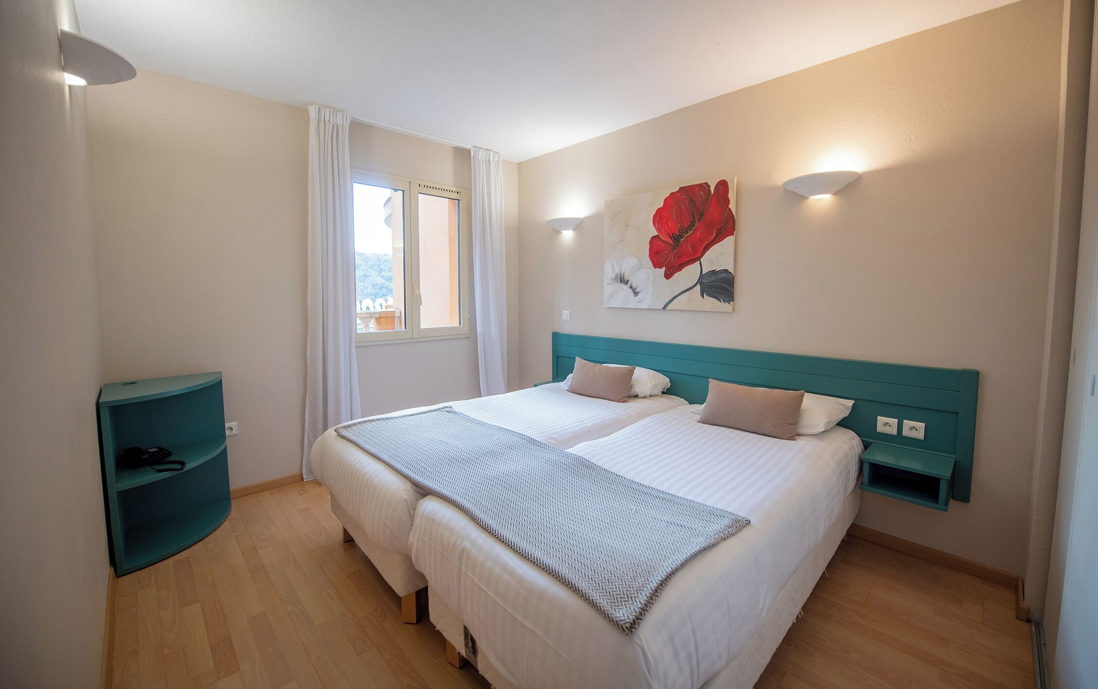 Un lit double avec une tête de lit de couleur turquoise, deux tables de chevet, une fenêtre avec des rideaux blancs et une œuvre d’art représentant des fleurs rouges, accrochée au mur.