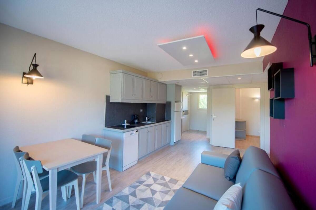 A modern kitchen with white cabinets, a dining table with chairs, and a sofa in a living area with a geometric rug.