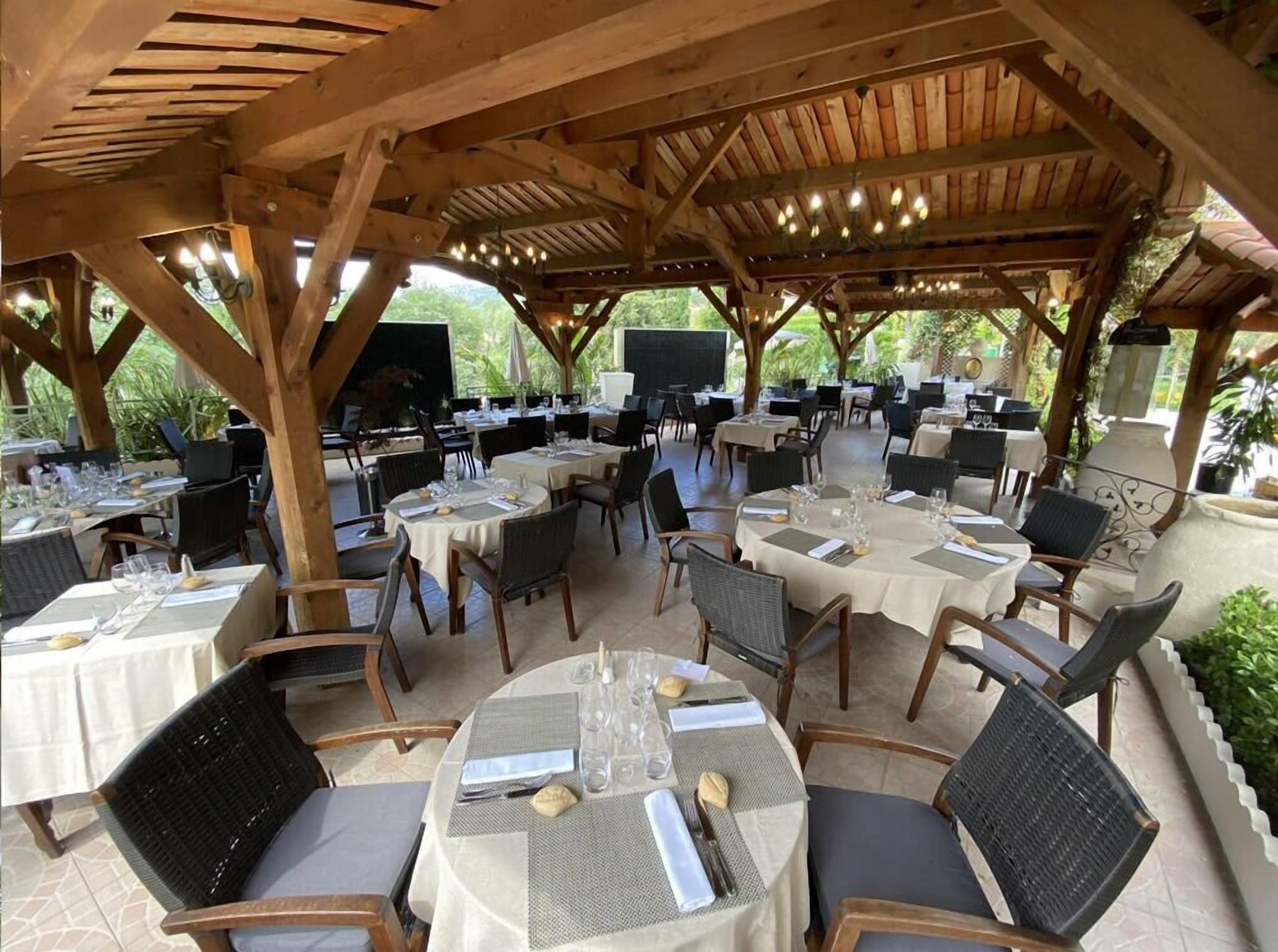 An outdoor dining area with round tables and chairs, covered by a wooden pergola with string lights.