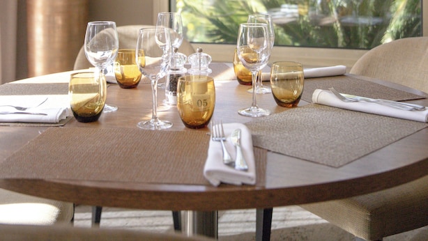 Une table de salle à manger ronde, agrémentée de six verres à vin et d’un grand vase, placée devant une fenêtre offrant une vue sur un paysage tropical.