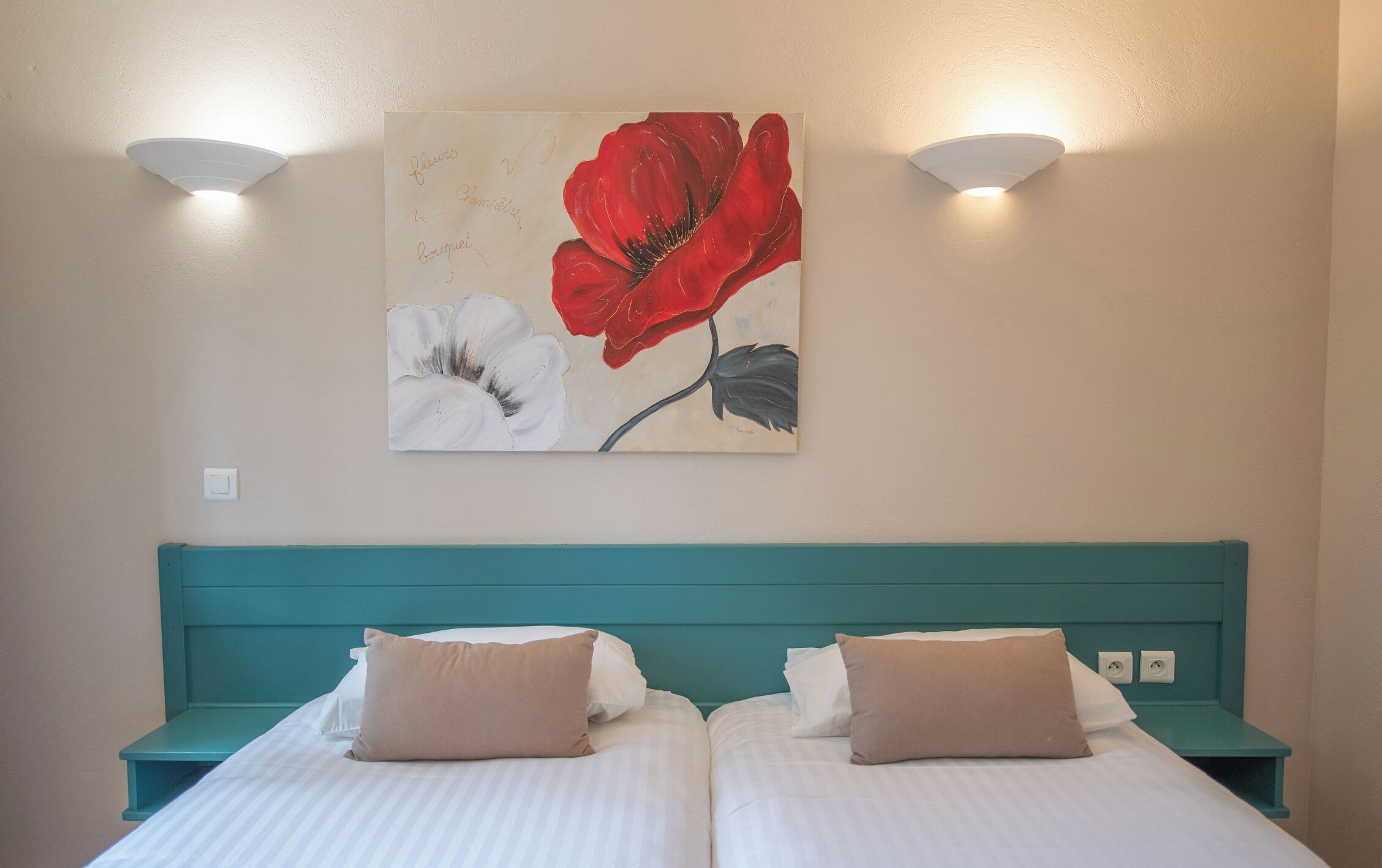 Een tweepersoonsbed met witte beddengoed en twee beige kussens, met aan weerszijden nachtkastjes in teal. Daarboven een wandlamp en een lijst met bloemistisch kunstwerk.