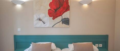 Een tweepersoonsbed met witte beddengoed en twee beige kussens, met aan weerszijden nachtkastjes in teal. Daarboven een wandlamp en een lijst met bloemistisch kunstwerk.