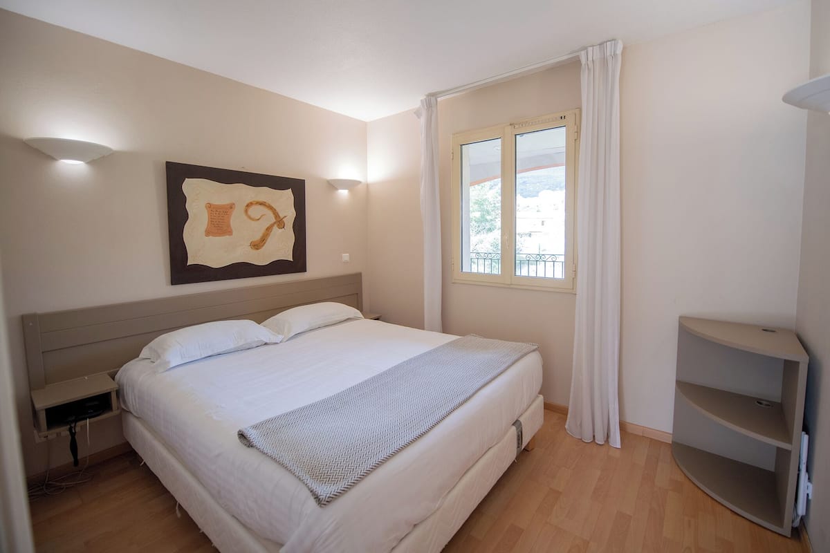 A neatly arranged bedroom with a bed, a bedside table, a window with curtains, and a wall-mounted artwork.