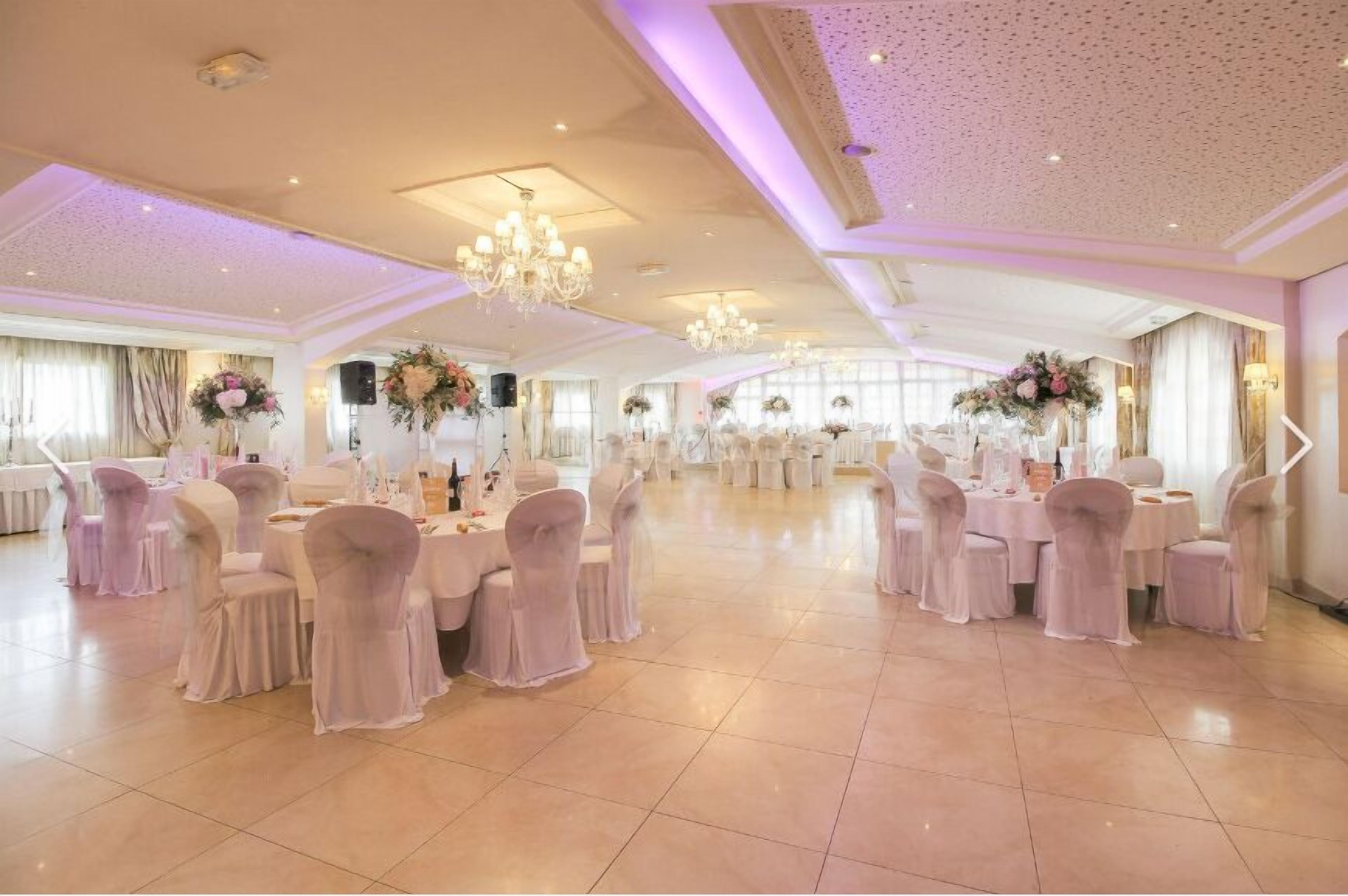 Uma ampla sala de banquetes com mesas redondas e cadeiras, lustres e centros de mesa com flores.
