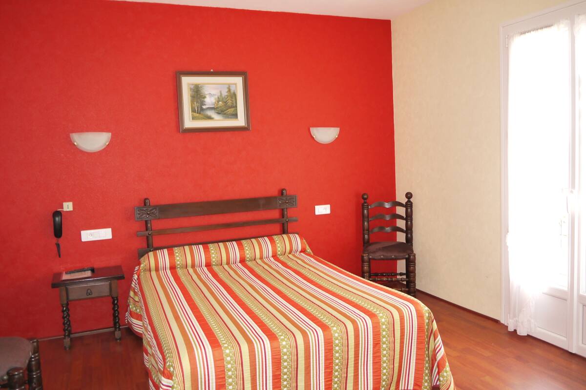 A bedroom with a red wall, a wooden bed frame, a striped bedspread, a nightstand, a chair, and a painting.