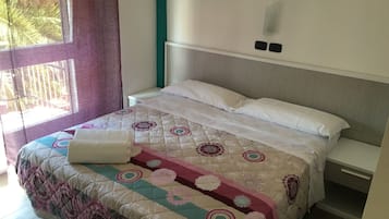 A neatly made bed with a patterned quilt and white pillows, a bedside table, and a window with curtains.