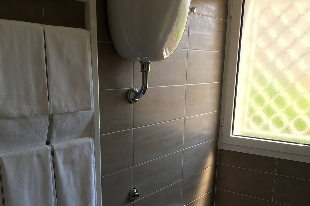 A bathroom with a toilet, a towel rack with multiple towels, a toilet paper holder, and a window with a view of greenery.