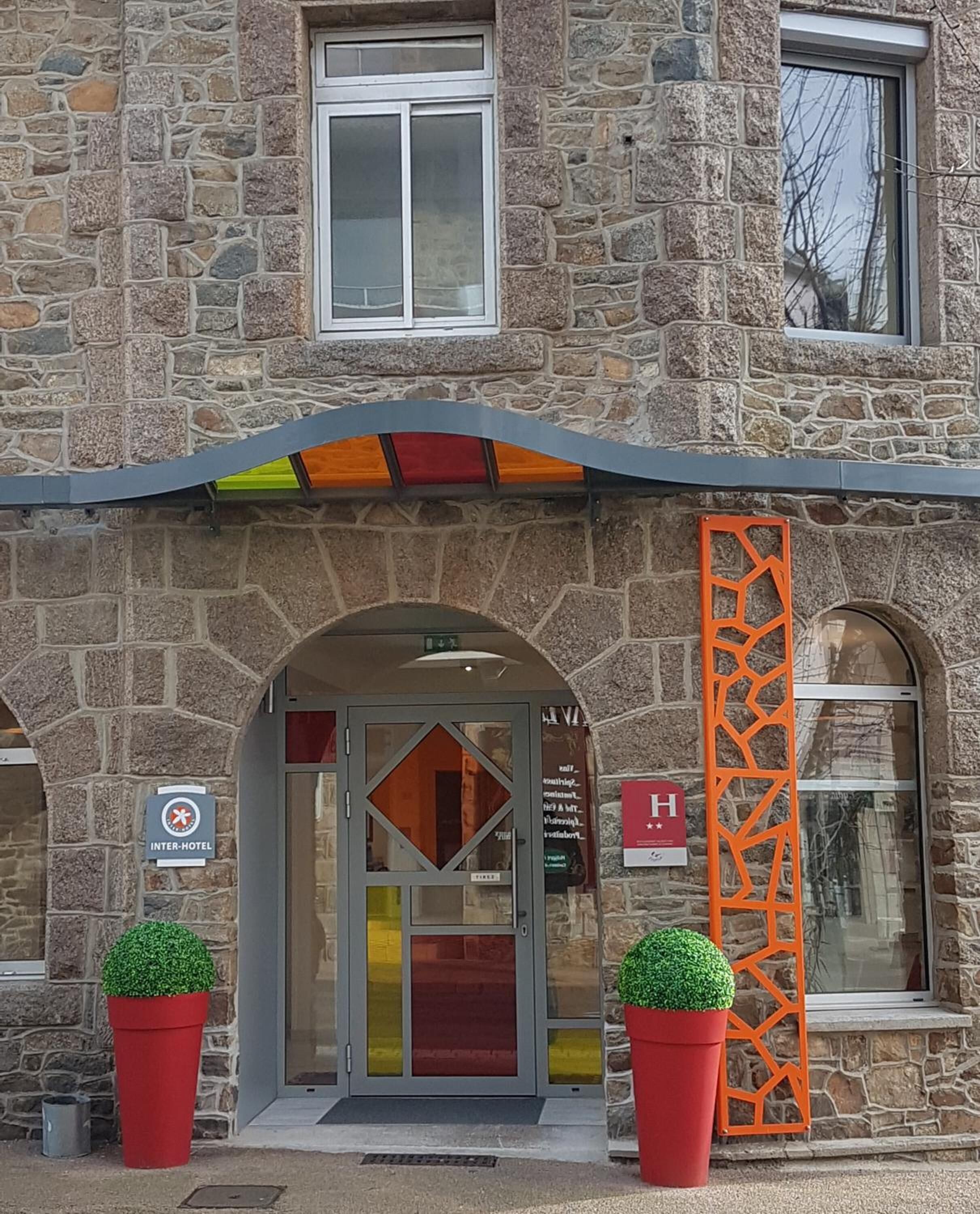Un bâtiment en pierre avec une entrée en verre, une enseigne indiquant qu’il s’agit d’un établissement proposant des services communs à plusieurs hôtels, et deux plantes en pot.