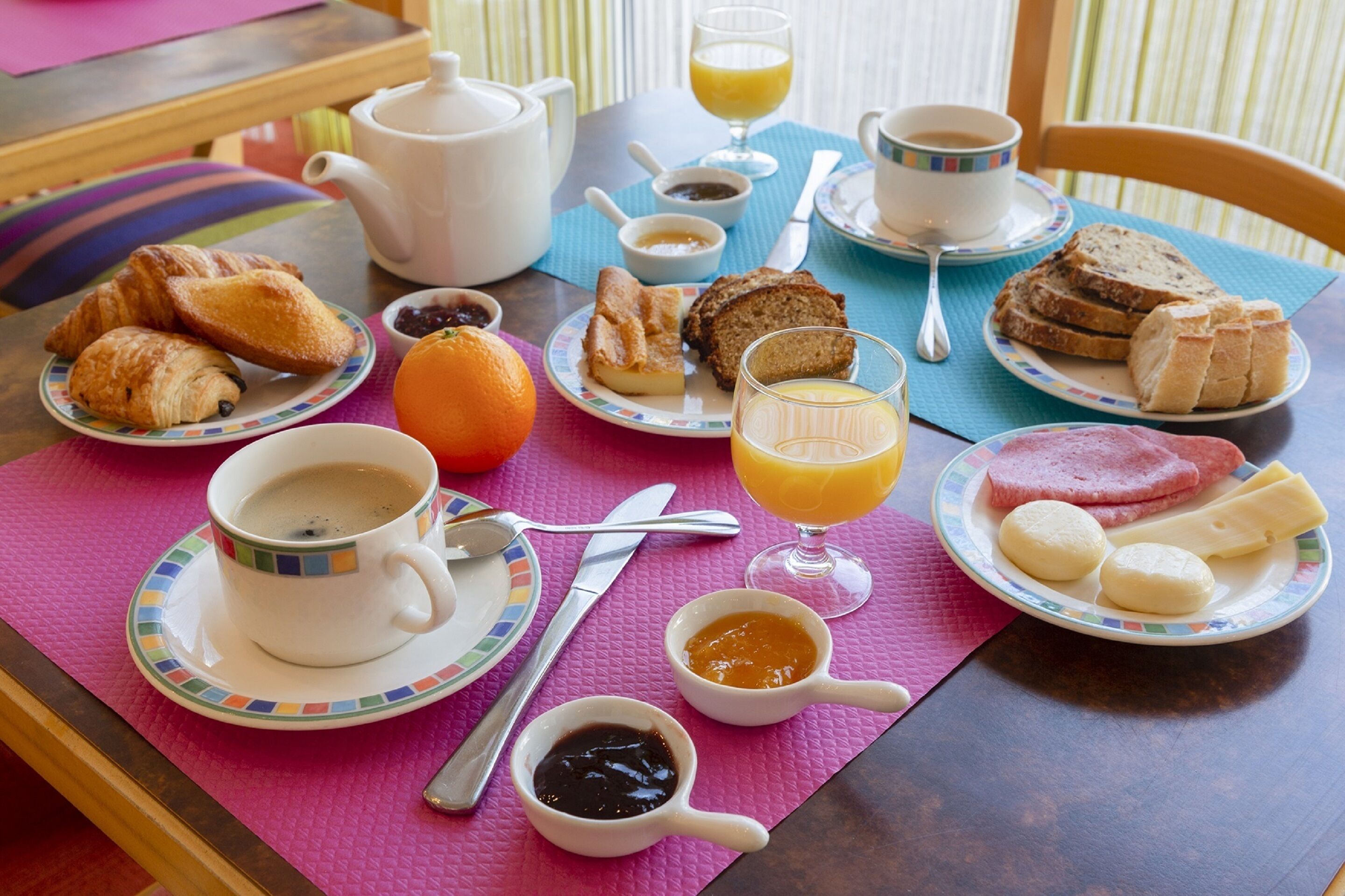 Una mesa de desayuno con croissants, pan, jamón, queso, jugo de naranja, café y té.