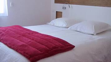 A neatly made bed with white linens and a red blanket, a wooden headboard, and a wall-mounted light fixture.