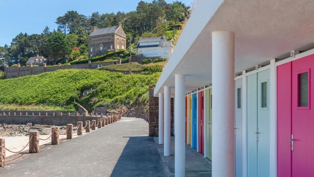 Um edifício costeiro com portas coloridas, uma parede de pedra e vista para uma colina com árvores e uma casa.