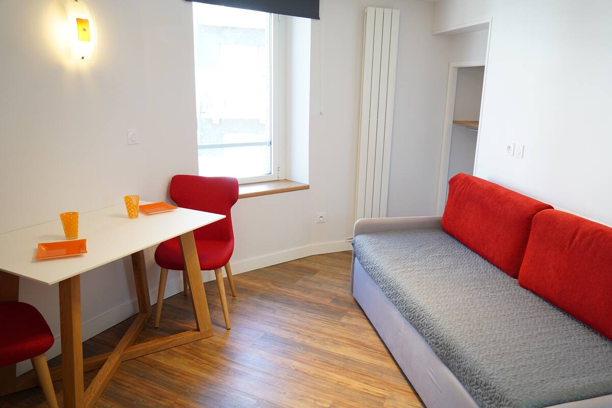 Une petite pièce bien éclairée, avec un plancher en bois, une table blanche et deux chaises rouges, un canapé gris avec des coussins rouges, et une fenêtre avec des stores.