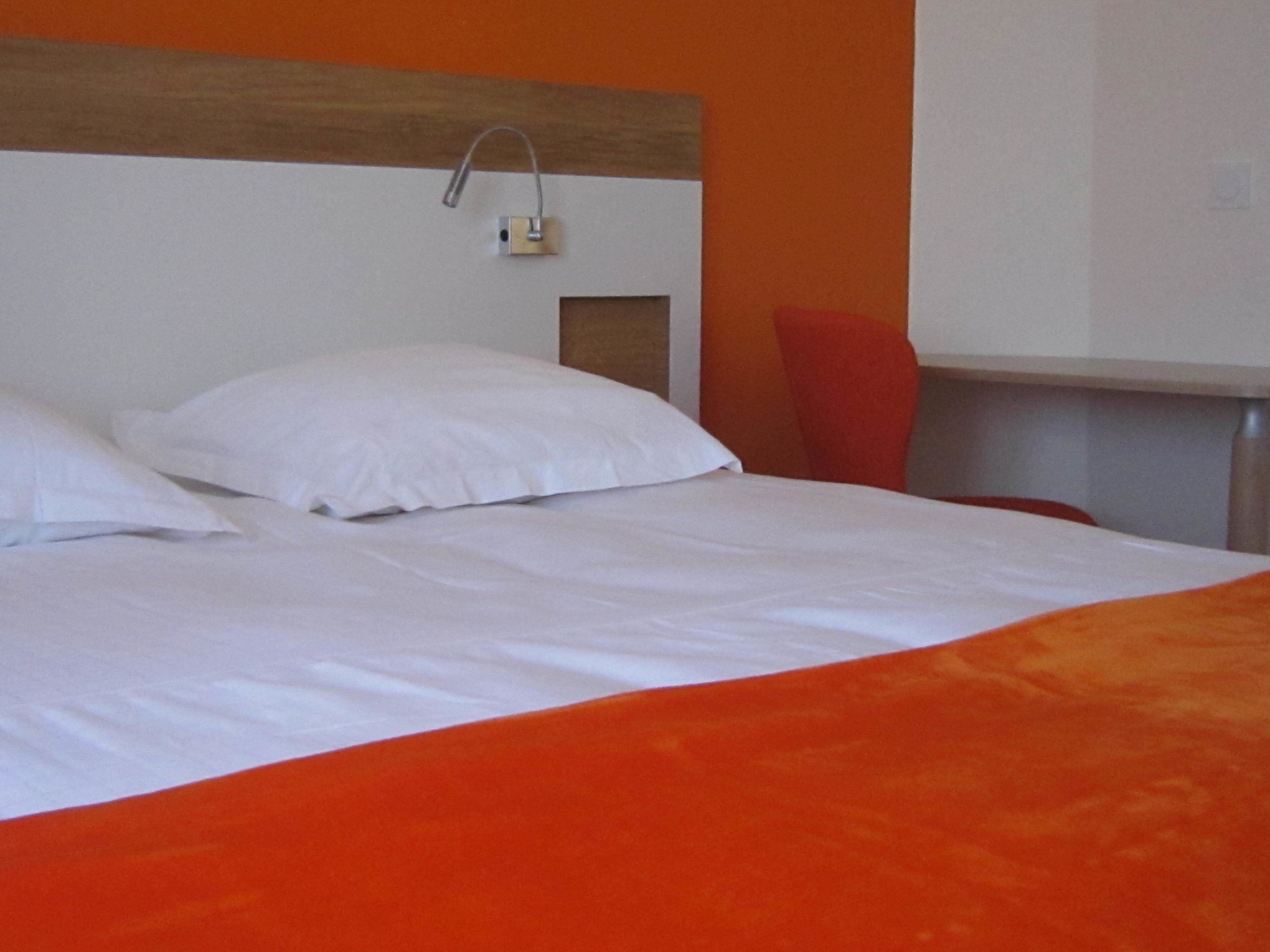 Cama com roupa de cama branca e um detalhe laranja, uma cadeira vermelha e uma cabeceira de madeira.
