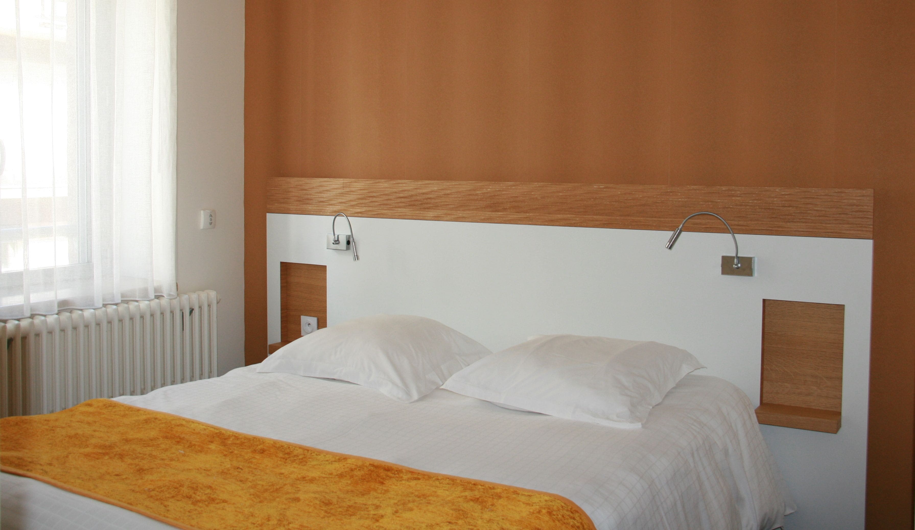 A hotel room with a bed, a wooden headboard, a window with sheer curtains, and a radiator.