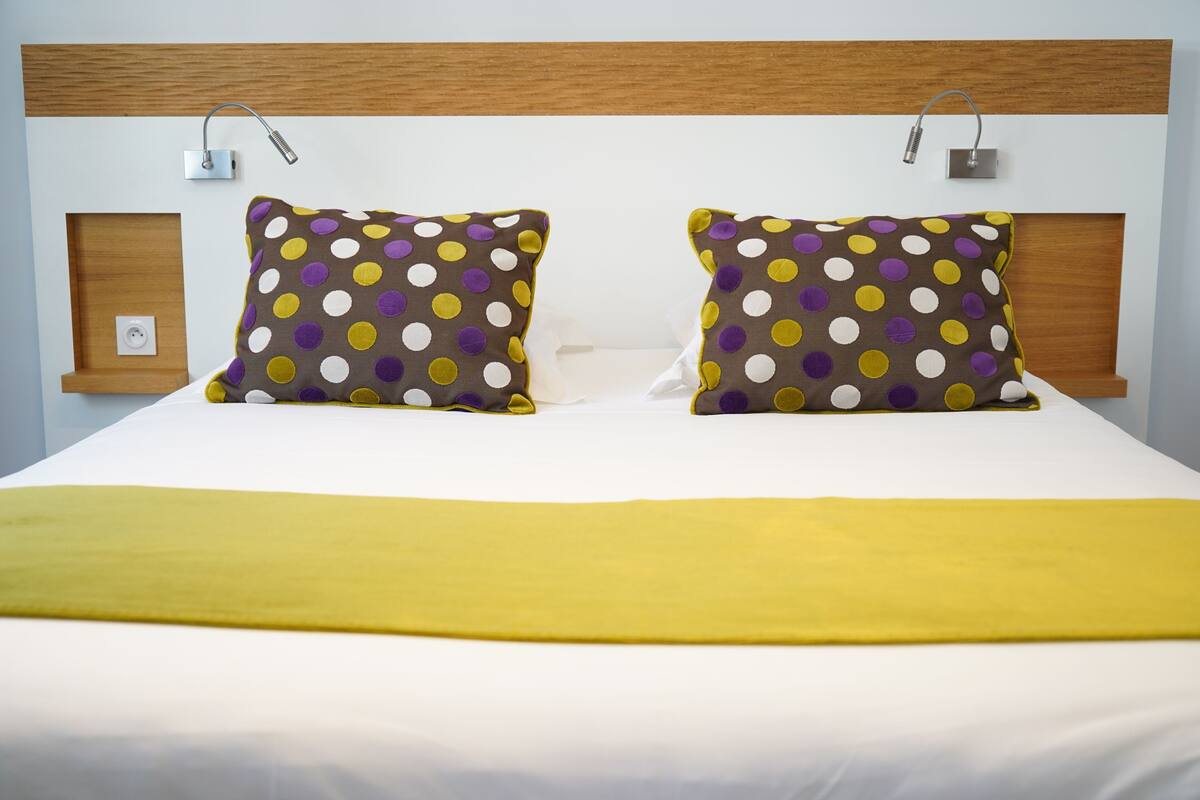 Un lit bien fait, agrémenté de deux coussins à pois et d’un plaid jaune.