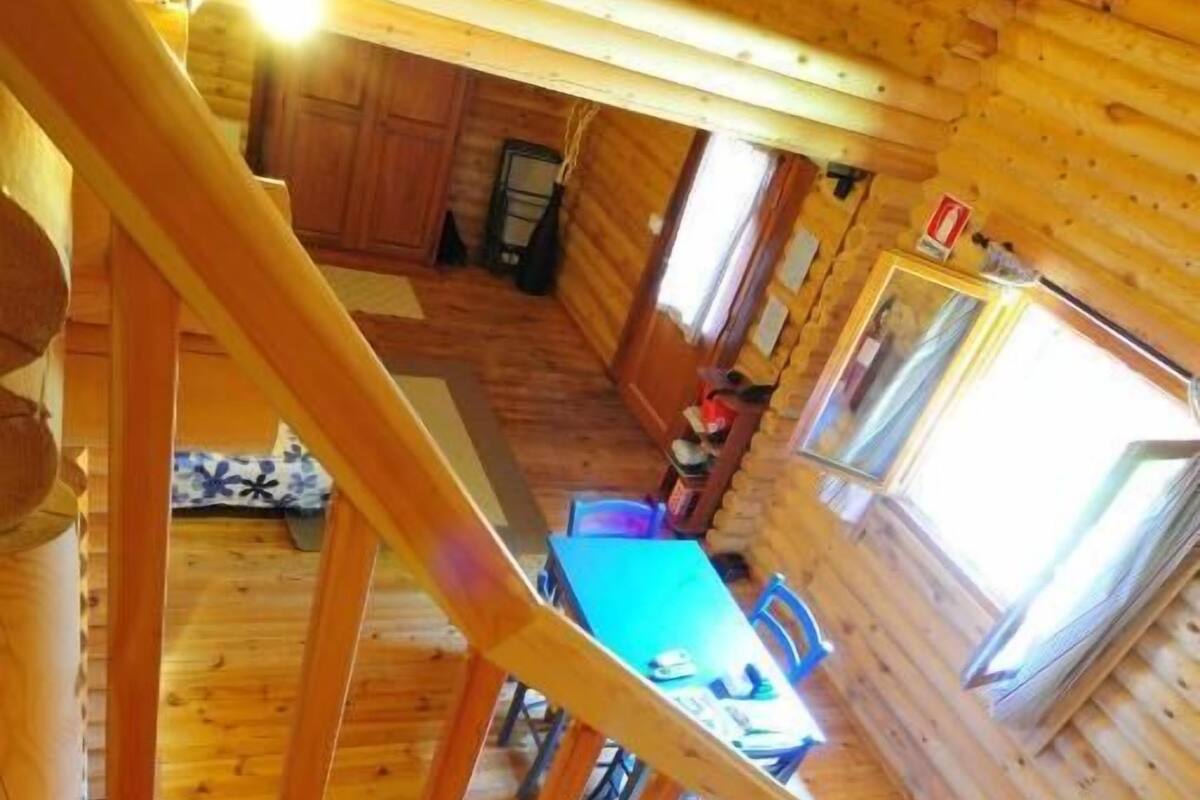 Interior de una cabaña de madera con comedor, escalera y vista al exterior.