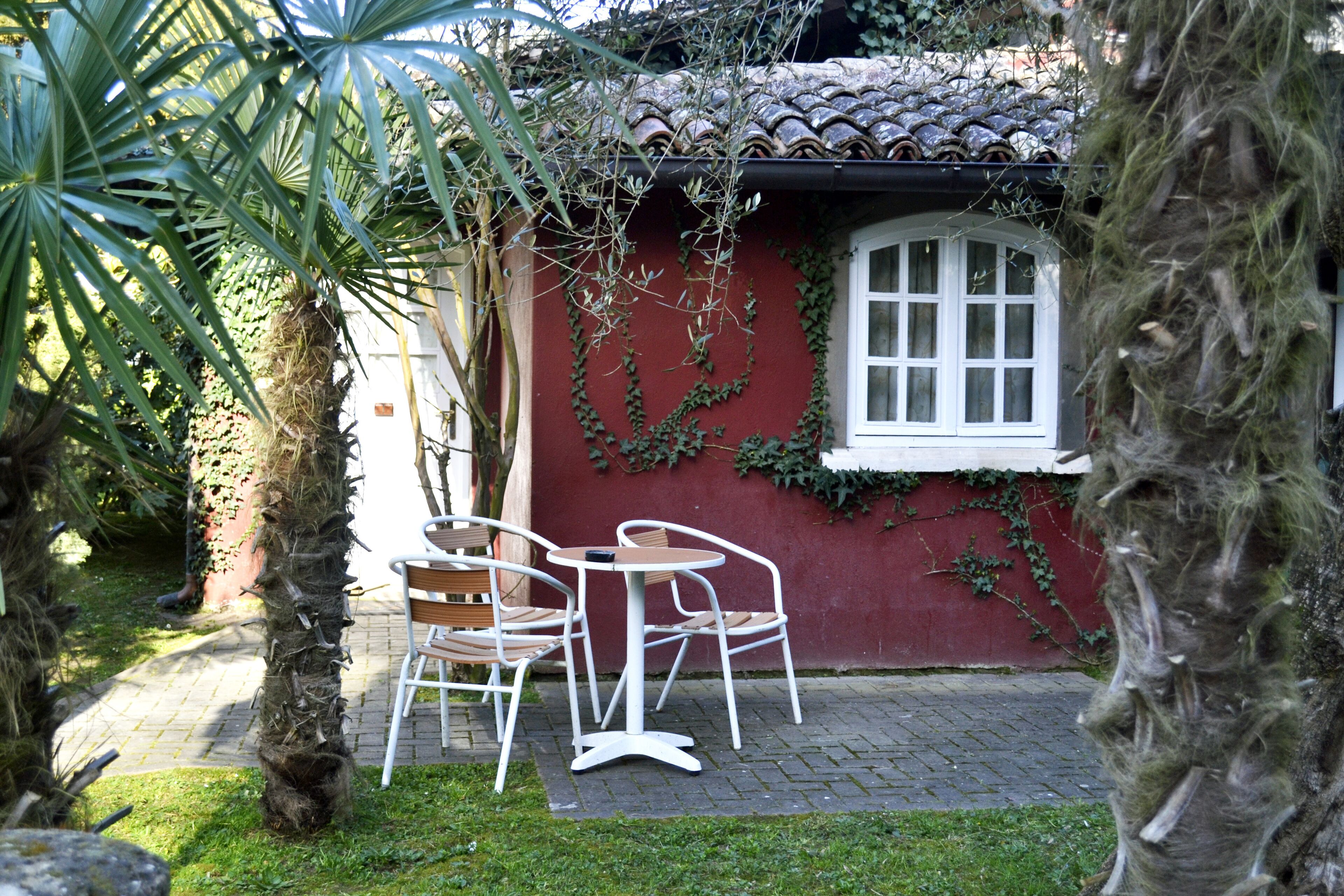 A small outdoor seating area with a table and chairs, surrounded by greenery and a red building with a tiled roof.