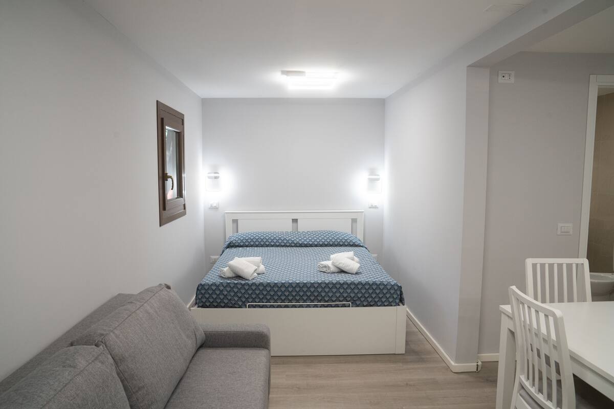 Un dormitorio pequeño y ordenado con cama, mesita de noche, silla y sofá.