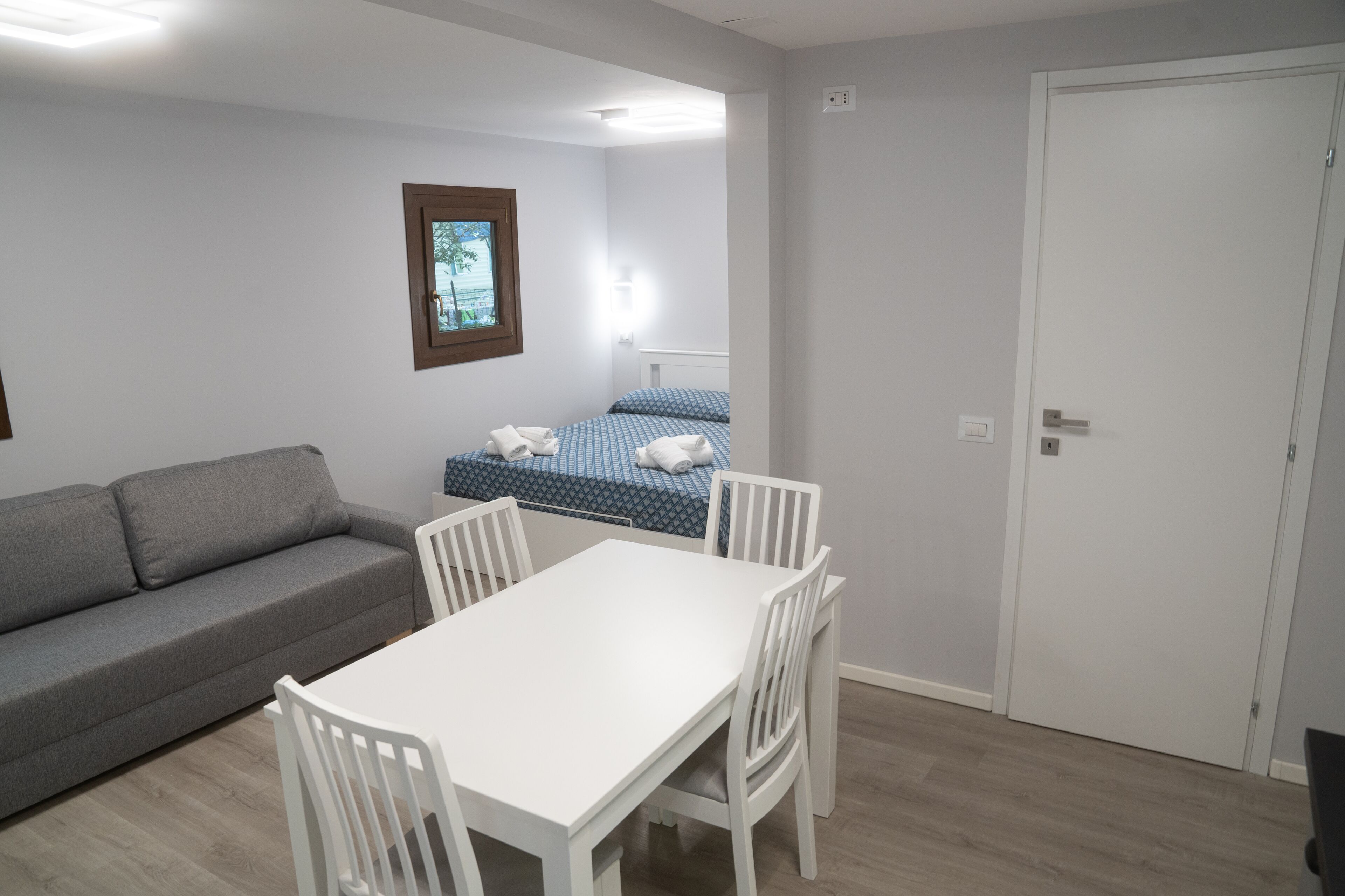 A small, well-lit room with a grey sofa, a white table with four chairs, a bed with blue bedding, and a door.