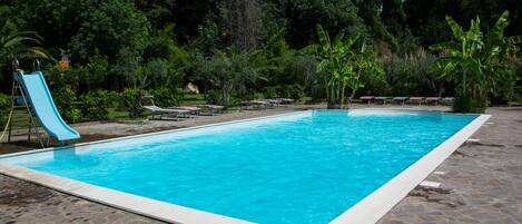 Piscina rectangular con solárium empedrado, rodeada de exuberante vegetación y un tobogán.
