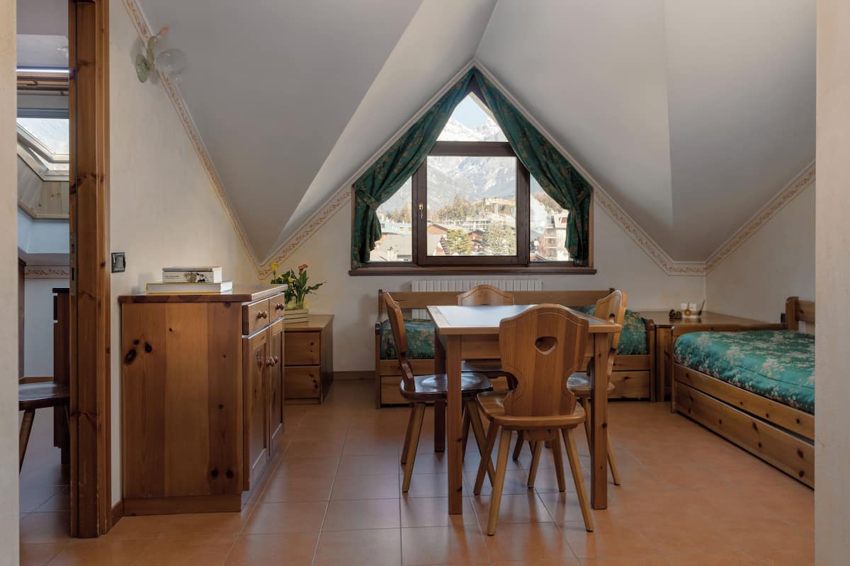 A cozy room with a wooden dining table, a bench, and a bed. There is a window with curtains and a view of mountains.