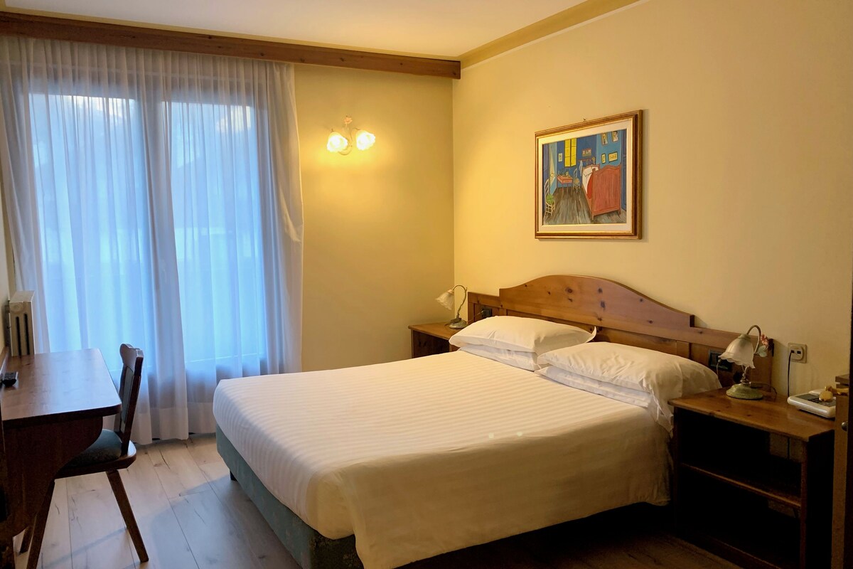 A hotel room with a bed, a desk, a chair, a nightstand, a lamp, and a painting on the wall.